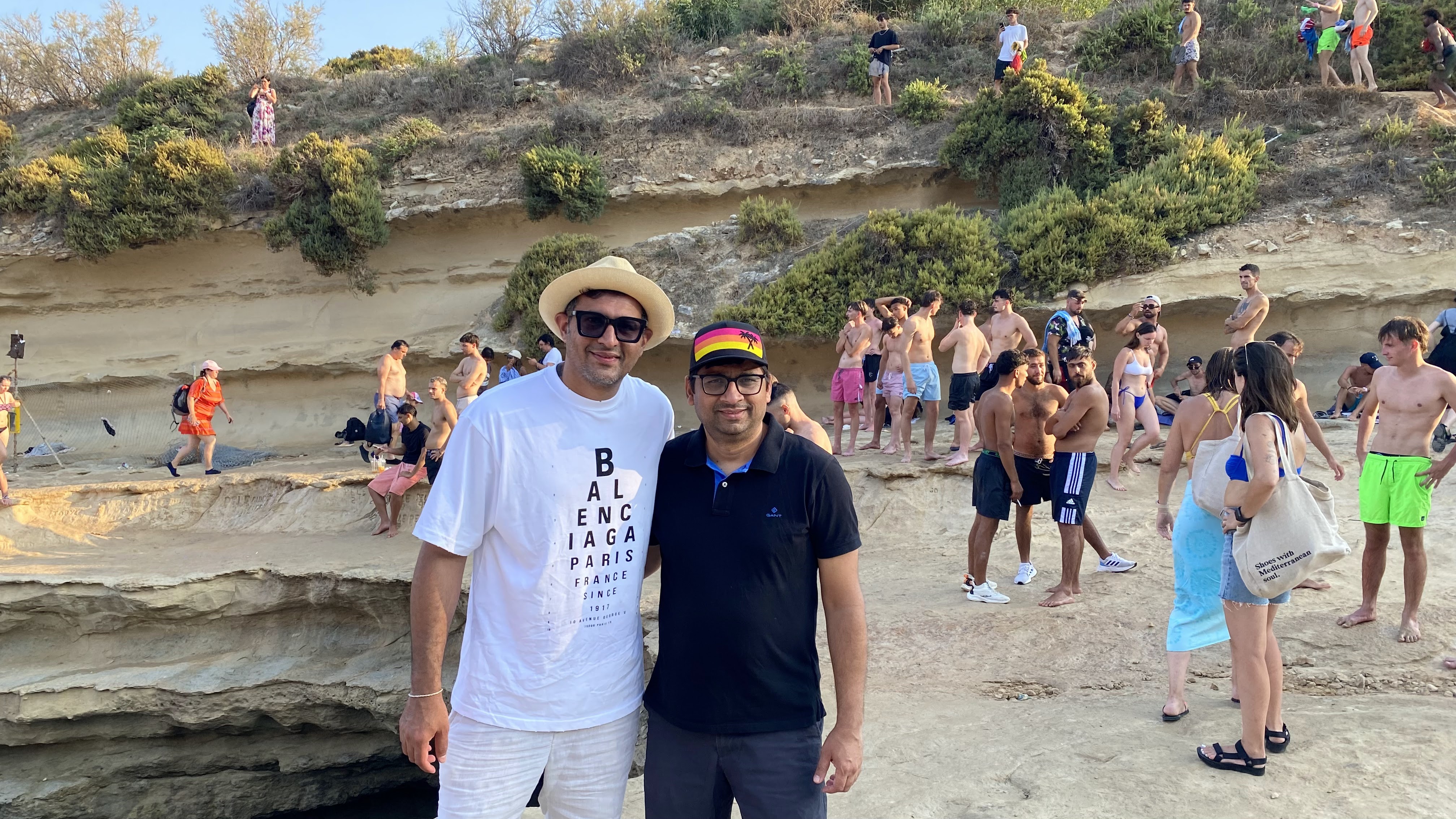 Two friends on a bustling rocky beach in Malta, surrounded by many beachgoers.